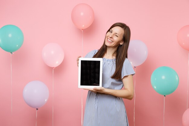 Portret van gelukkig lachende jonge vrouw in blauwe jurk met tablet pc-computer met leeg leeg scherm knipperend op pastel roze achtergrond met kleurrijke luchtballonnen. Verjaardagsfeestje concept.