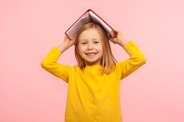 Portret van gelukkig lui meisje dat hoofd bedekt met boek en glimlacht naar camera ongehoorzaam kind dat plezier heeft met gek rond in plaats daarvan studeren les binnen studio opname geïsoleerd op roze achtergrond