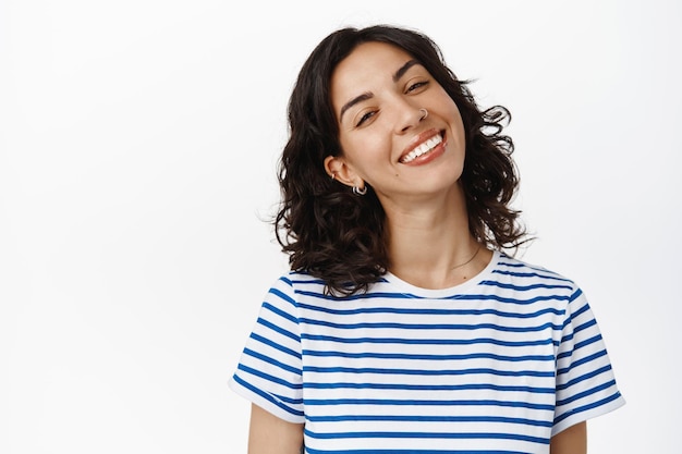 Portret van gelukkig mooi meisje met donker krullend haar, doorboorde neus, lachende witte tanden, positief en vrolijk kijkend, genietend van het leven, staande in zomer t-shirt tegen witte achtergrond.