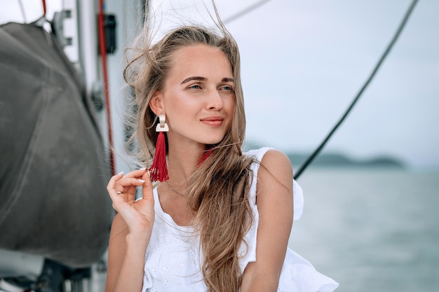 Portret van gelukkig mooi meisje met witte jurk, lange rode oorbellen en lang krullend blond haar staande op jacht in de zomer. camera kijken met een brede glimlach. detailopname