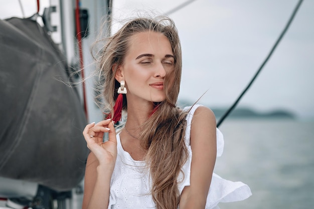 Portret van gelukkig mooi meisje met witte jurk, lange rode oorbellen en lang krullend blond haar staande op jacht in de zomer. camera kijken met een brede glimlach. detailopname