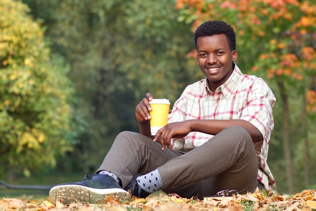 Portret van gelukkig positieve zwarte Afro-Amerikaanse jonge knappe man zittend op het gras in gouden herfst park, glimlachen, een warme drank thee of koffie drinken uit plastic beker