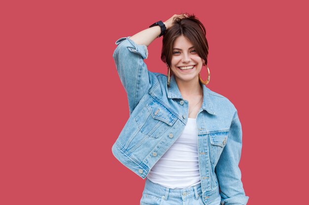 Portret van gelukkig tevreden mooie brunette jonge vrouw met make-up in denim casual stijl staan, poseren en kijken naar camera met brede glimlach. indoor studio opname, geïsoleerd op rode achtergrond.