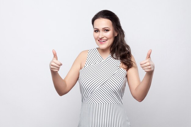 Portret van gelukkig tevreden mooie jonge brunette vrouw met make-up en gestreepte jurk staan, toothy glimlachen en kijken naar camera met duimen omhoog. indoor studio opname, geïsoleerd op een grijze achtergrond.