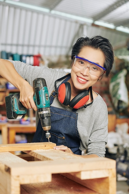 Portret van gelukkig timmerman in beschermende bril met behulp van boor bij het maken van houten meubels in werkplaats