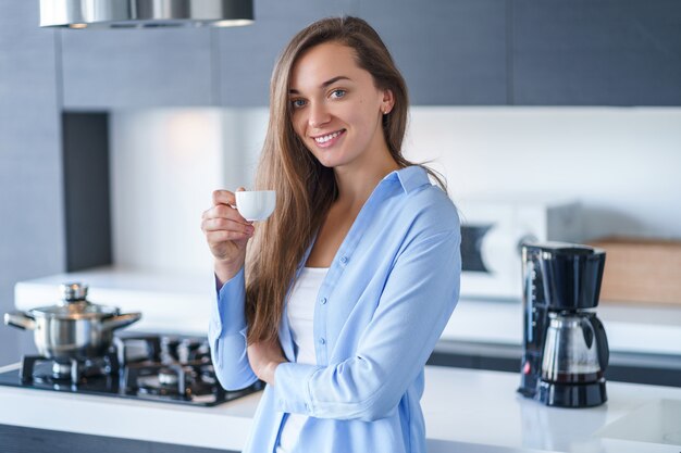 Portret van gelukkige aantrekkelijke vrouw die en van hete verse aromatische koffie drinken genieten na het brouwen van koffie thuis gebruikend koffiezetapparaat in de keuken