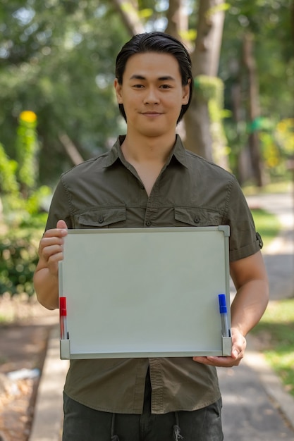 Portret van gelukkige Aziatische jonge mens die zich in het park bevindt en leeg uithangbord toont