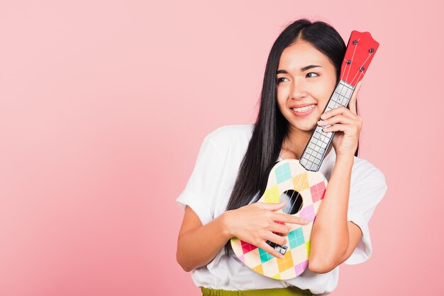 Portret van gelukkige Aziatische mooie jonge vrouw tiener zelfverzekerd lachend gezicht houden akoestische ukulele gitaar, vrouwelijke spelen Hawaiiaanse kleine gitaar, studio opname geïsoleerd op roze achtergrond, met kopie ruimte