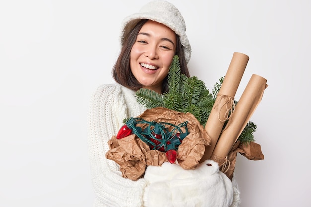 Portret van gelukkige Aziatische vrouw bereidt zich voor op vakantie glimlacht breed draagt hoed gebreide trui wanten omarmt vuren takken met papier voor verpakking en retro slinger versiert huis voor Nieuwjaar