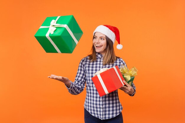 Portret van gelukkige brunette jonge vrouw in kerstmuts en geruit hemd staande geschenkdozen in de lucht te gooien of cadeautjes te vangen kerstinkopen binnen studio opname geïsoleerd op oranje achtergrond