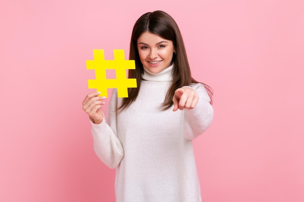 Portret van gelukkige brunette vrouw met brede glimlach wijzend op camera met gele hash symbool dragen witte casual stijl trui Indoor studio opname geïsoleerd op roze achtergrond