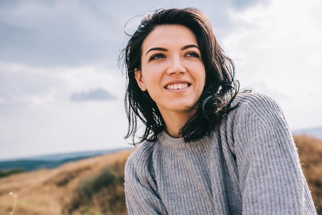 Portret van gelukkige en grappige brunette vrouw glimlachend en wegkijkend tegen natuurweide en bewolkte achtergrond met winderig haar Mensen reizen en lifestyle concept Cover mood