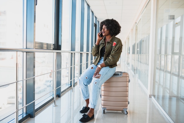 Portret van gelukkige jonge Afro-Amerikaanse vrouw zittend op een koffer en praten met een mobiele telefoon op het station