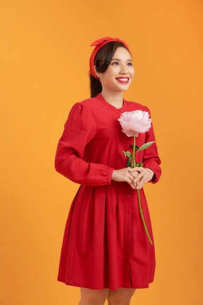 Portret van gelukkige jonge Aziatische vrouw die pioenbloem over oranje achtergrond houdt