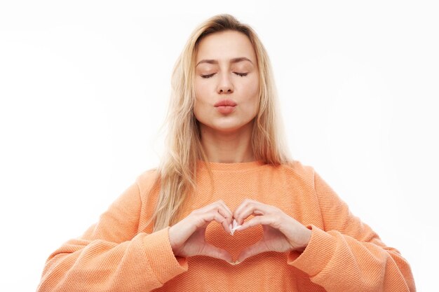 Portret van gelukkige jonge blonde vrouw maakt een hartgebaar en vraagt om mijn valentijn te zijn. Vriendin houdt de handen op de borst en voelt zich dankbaar geïsoleerd op een witte studioachtergrond