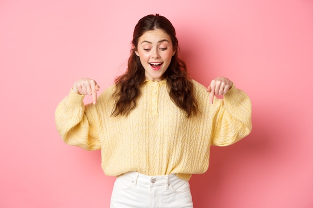 Portret van gelukkige jonge donkerbruine vrouw die promo-aanbieding bekijkt, en met verbaasd gezicht naar beneden kijkt, die zich over roze muur bevindt.