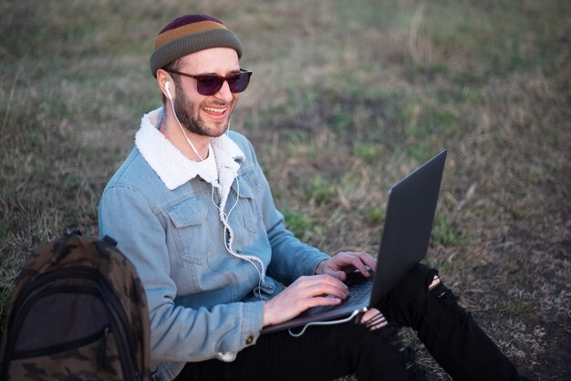 Portret van gelukkige jonge man in park, zittend met laptop op benen in de buurt van rugzak Gekleed in jeans jasje met bril en hoed