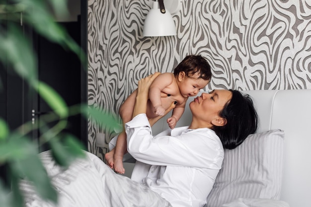 Portret van gelukkige jonge moeder zoenen met liefde schattige dochtertje in slaapkamer wazig groene planten op voorgrond Rusten in grijs bed met naakte baby Concept van moederlijke genegenheid en kinderopvang