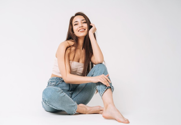 Portret van gelukkige jonge mooie aziatische vrouw met donker lang haar in spijkerbroek zittend op een witte achtergrond geïsoleerd