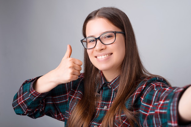 Portret van gelukkige jonge vrouw die selfie en duim over witte muur tonen nemen