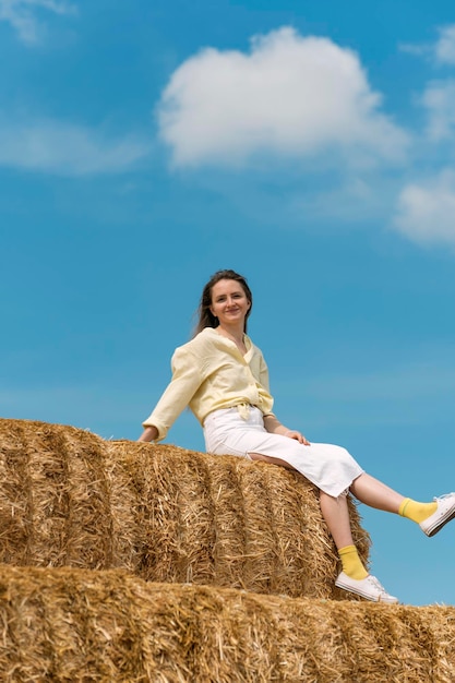 Portret van gelukkige jonge vrouw zittend op hoge hooiberg op blauwe hemelachtergrond Glimlachende vrouw in zomervakantie Harvesting