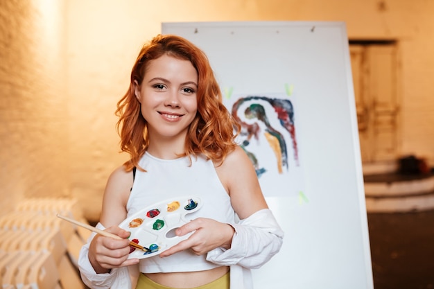 Foto portret van gelukkige jonge vrouwenschilder met rood haar die zich op leeg canvas in kunstenaarsatelier bevinden.