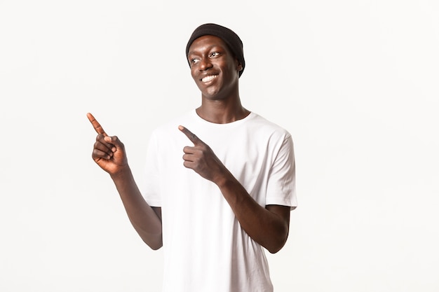 Portret van gelukkige knappe Afro-Amerikaanse jonge man, met beanie en t-shirt, glimlachend tevreden en kijkend, linker bovenhoek wijzend