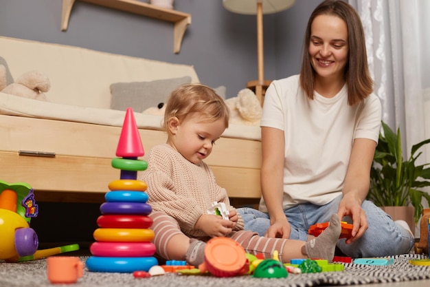 Portret van gelukkige moeder interactie met peuter babymeisje in de kinderkamer zittend op de vloer in de woonkamer spelen met kleurrijk speelgoed tijd samen doorbrengen kind onderwijs