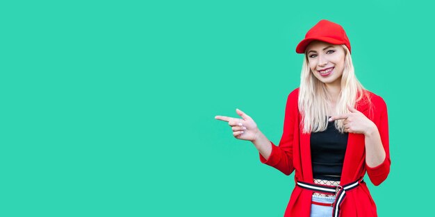 Portret van gelukkige mooie blonde jonge hipster vrouw in rode blouse en pet, staand, wijzend op copyspace en kijkend naar camera met brede glimlach. indoor studio opname, geïsoleerd op groene achtergrond.