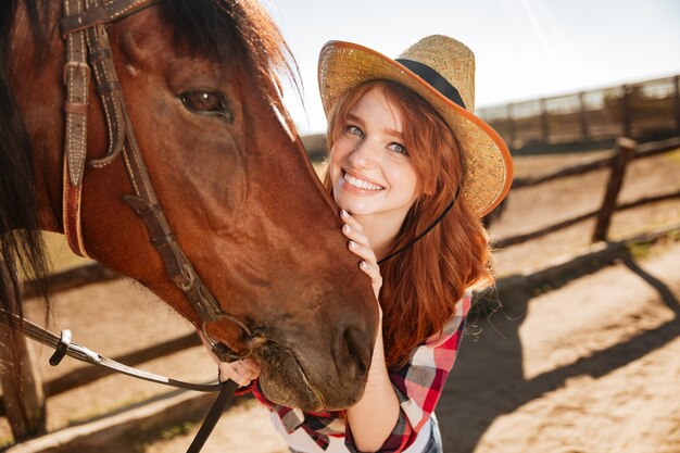 Portret van gelukkige mooie jonge vrouwenveedrijfster in hoed met haar paard op boerderij