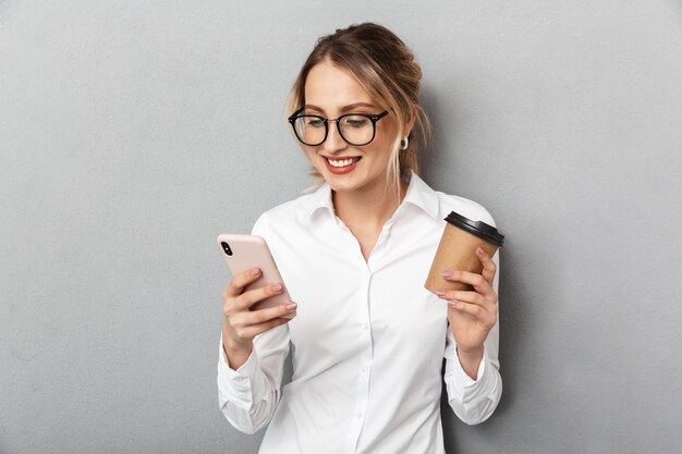 Portret van gelukkige onderneemster die glazen draagt die smartphone houdt en koffie drinkt op kantoor, geïsoleerd