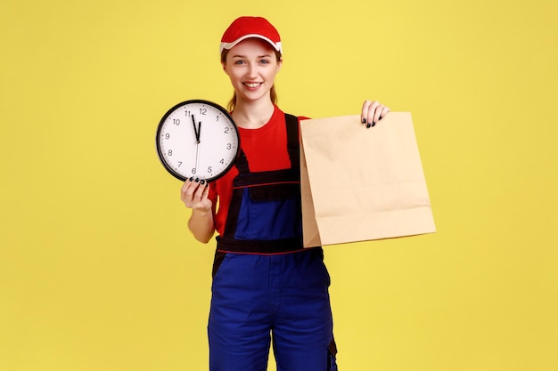 Portret van gelukkige positieve koeriersvrouw met pakket en wandklok snelle bezorgservice die op tijd levert met overall en rode dop Indoor studio-opname geïsoleerd op gele achtergrond