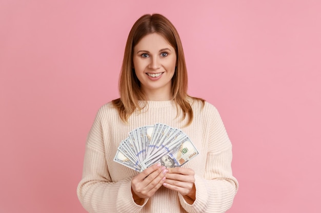 Portret van gelukkige rijke blonde vrouw met grote fan van dollar biljetten kijken camera met positieve uitdrukking dragen witte trui Indoor studio shot geïsoleerd op roze achtergrond