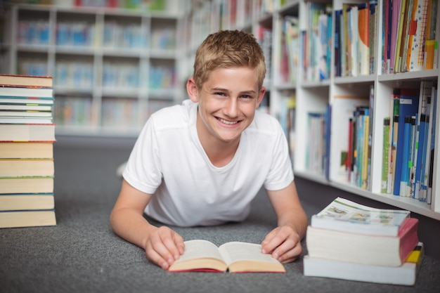 Portret van gelukkige schooljongen die op vloer met boeken in bibliotheek ligt