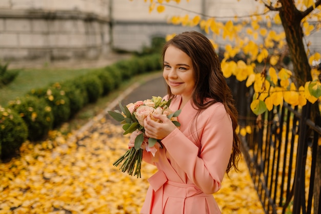 Portret van gelukkige sensuele jonge donkerbruine bruid in roze huwelijkskleding en status in de herfstpark dichtbij uitstekende kathedraal en het houden van huwelijksboeket.