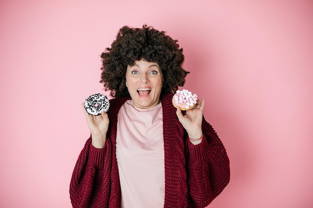 Portret van gelukkige vrouw die beetwen twee donutes op roze achtergrond kiest. Afro kapsel, humor