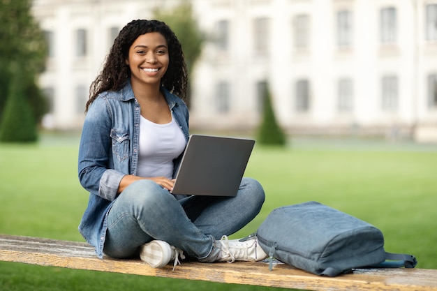 Portret van gelukkige zwarte vrouwelijke student met laptop zittend op een bankje buitenshuis