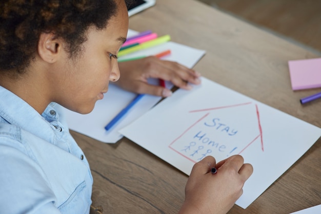 Portret van gemengd ras tiener schoolmeisje tekening blijf thuis inscriptie op papier met kleurrijke markeringen