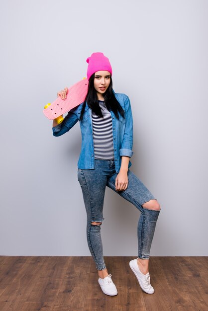 Portret van gemiddelde lengte van jonge glimlachende vrouw in jeans en roze hoed die kleurrijk skateboard op haar schouder houden tegen grijze ruimte