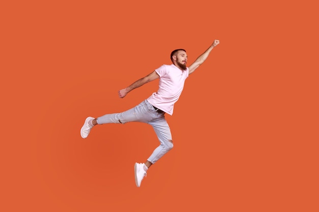 Portret van gemotiveerde zelfverzekerde bebaarde man die in de lucht vliegt als superheld met opgeheven hand, superkracht voelt en een roze T-shirt draagt. Indoor studio opname geïsoleerd op een oranje achtergrond.