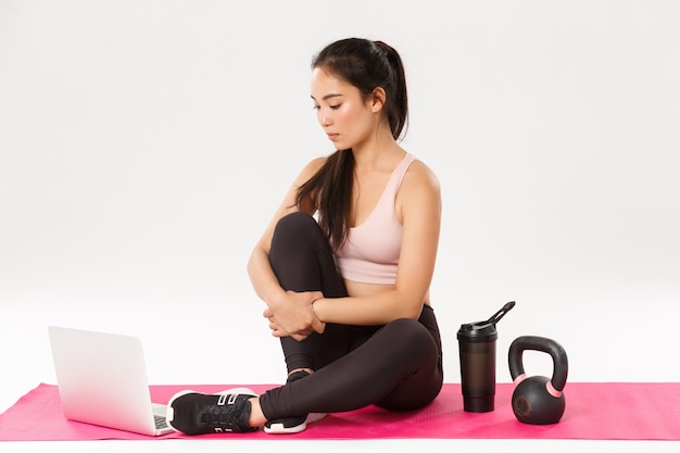 Portret van gerichte schattige Aziatische fitness meisje training vanuit huis, zittend op een rubberen mat met sportartikelen, laptop kijken, online coach bellen, oefeningsvideo op internet kijken.