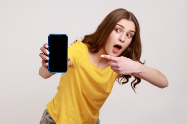 Portret van geschokt jonge vrouw in gele casual tshirt staande telefoon te houden en wijzende vinger met verrast gezicht en open mond Indoor studio opname geïsoleerd op grijze achtergrond