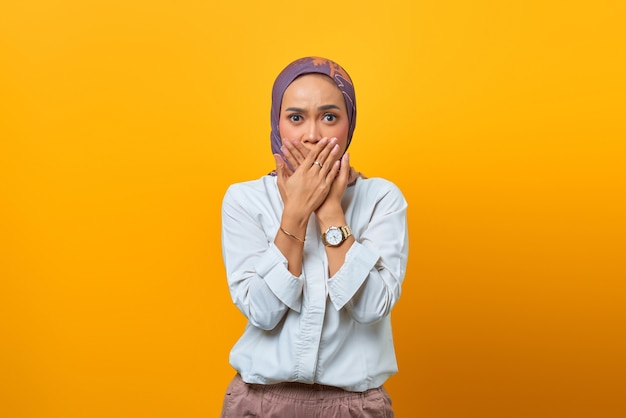 Portret van geschokte Aziatische vrouw terwijl bedekte mond met handen over gele achtergrond