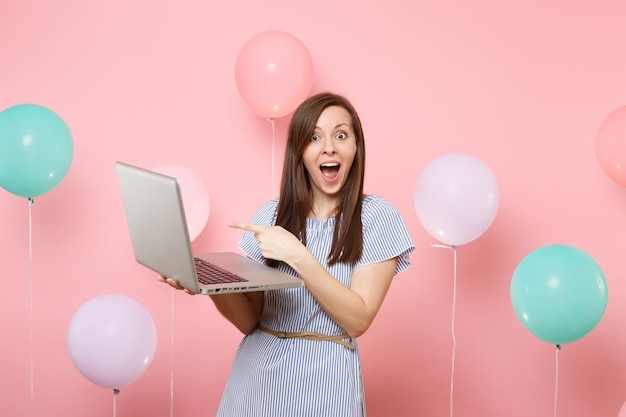 Portret van geschokte jonge vrouw in blauwe jurk met wijzende wijsvinger op laptop pc-computer op roze achtergrond met kleurrijke luchtballonnen. Verjaardag vakantie partij mensen oprechte emoties concept.