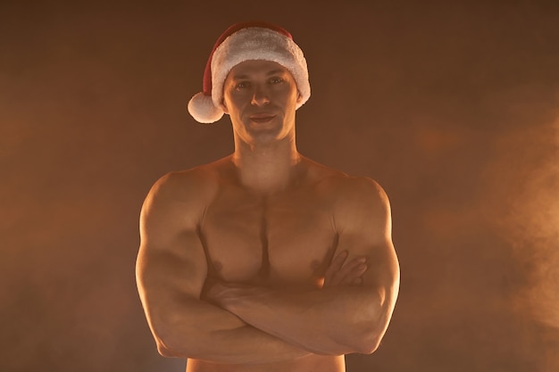 Portret van gespierde man met kerst kerstmuts, gevouwen handen en glimlach op rokerige achtergrond Macho Shirtless naakte torso stripper hete sexy kerstman Verleidelijke man voor vakantie