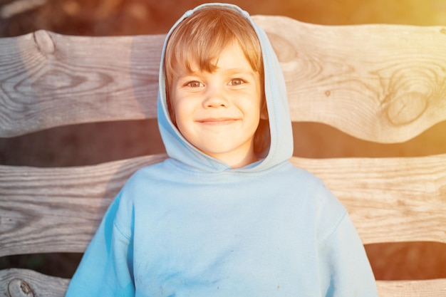 Portret van gezicht gelukkig lachend openhartige vijfjarige jongen jongen in blauwe hoodie in een kap op de achtergrond van houten planken in de aard van de zomer in een gouden uur bij zonsondergang homeschool en reizen flare