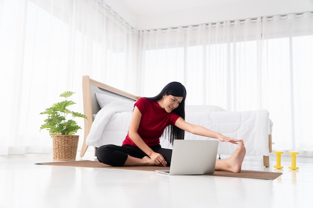 Portret van gezonde jonge Aziatische vrouw beoefenen van yoga oefeningen in de slaapkamer zitten en online leren op laptop thuis.