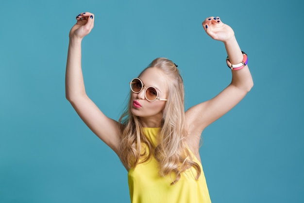 Portret van glamoureuze mooie blonde vrouw in zonnebril en geel shirt dansen op blauwe achtergrond. Zorgeloze zomer.