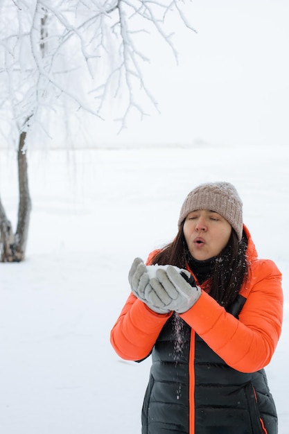 Portret van glanzende vrouw van middelbare leeftijd sneeuw uit handen blazen in handschoenen met twijgen bedekt met sneeuw op de achtergrond op wandeling in het besneeuwde bos Magische wintertijd vol witte kleur