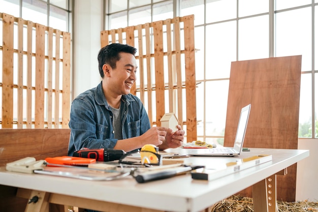 Portret van glimlach Aziatische mannelijke vader die een klein houten huis bouwt en bij elkaar houdt in de houtbewerkingsstudio thuis met het meetinstrument en werkgereedschap op tafel